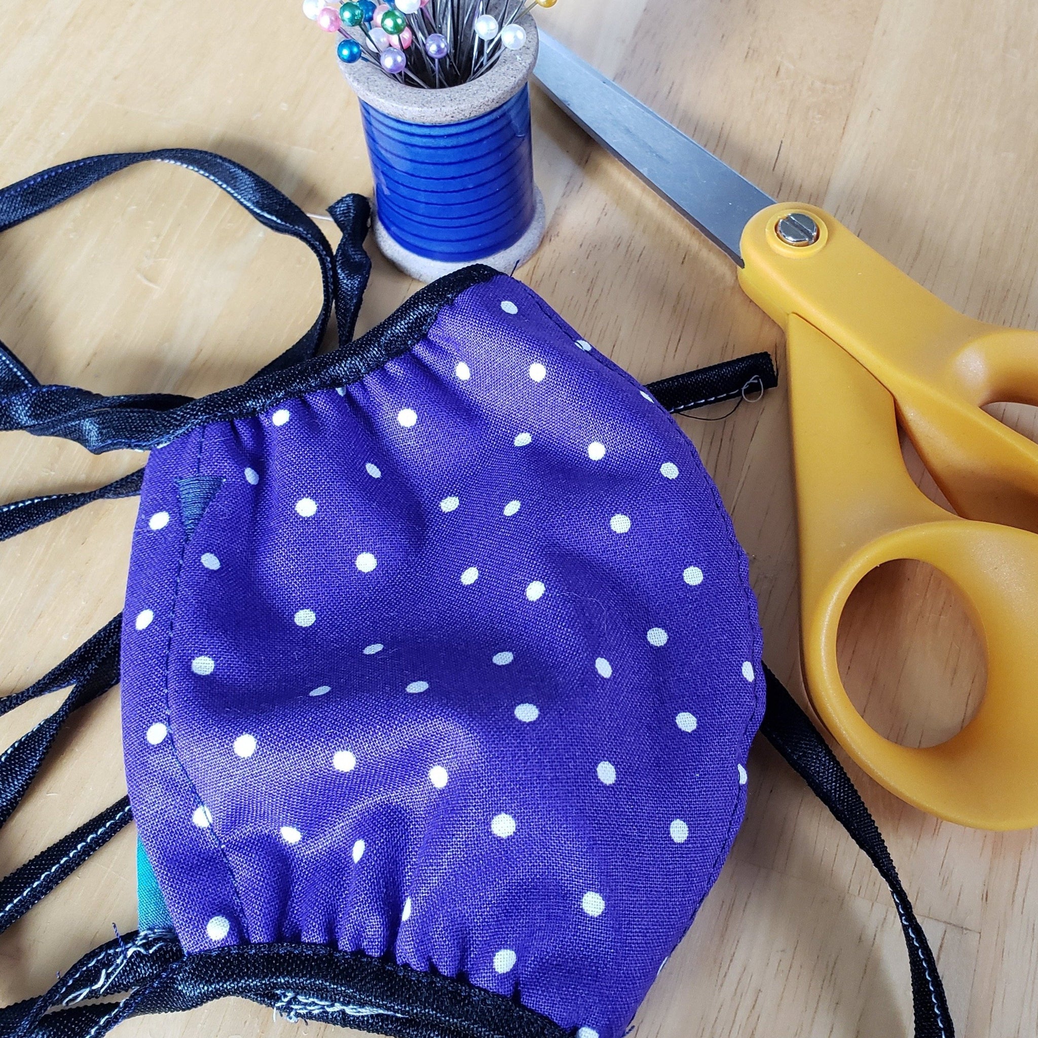 Purple polka dot face mask on table, shown with pins and scissors.